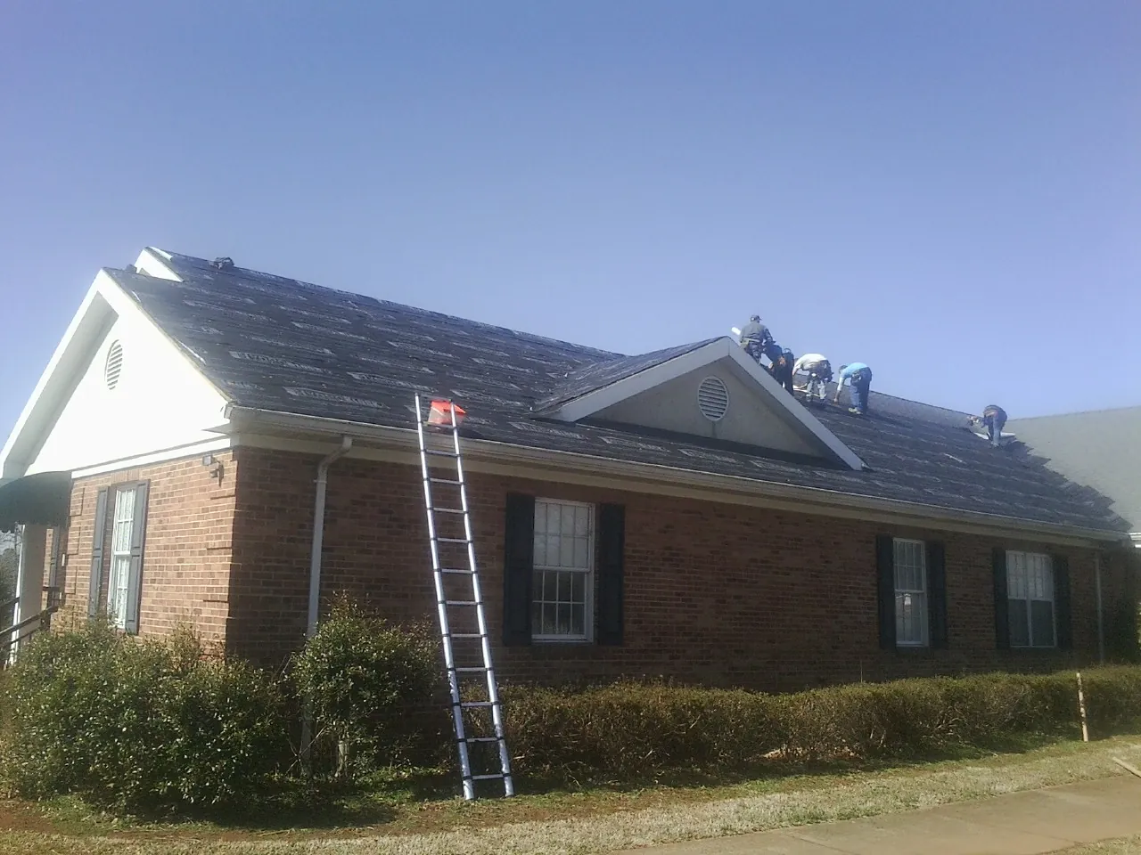 Crew tearing shingles off a brick ranch roof with a ladder set — Metal Roof Repair work by our crew in Guide Rock