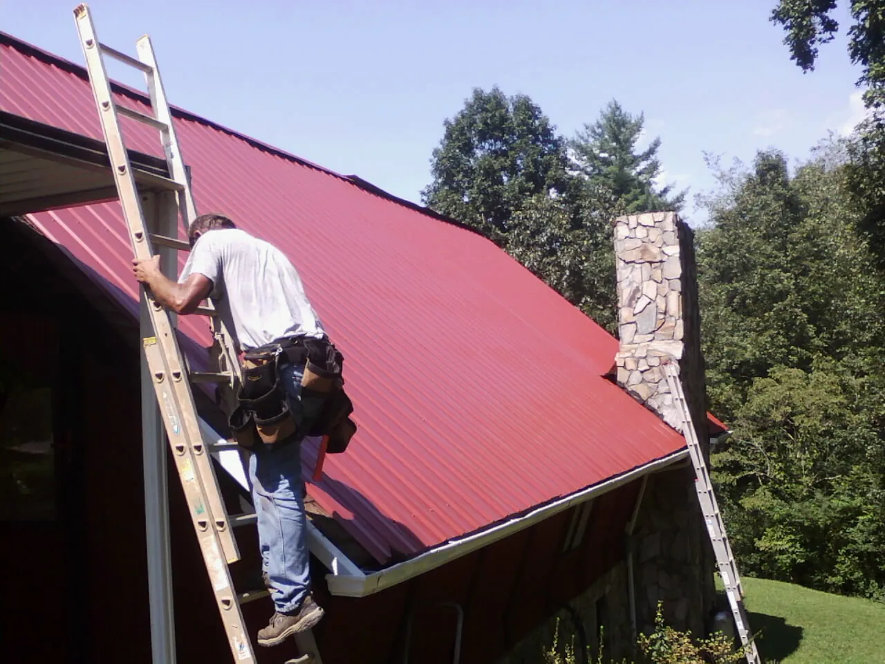 Roofer on a ladder beside a red metal roof and stone chimney — Soffit & Fascia Repair by Roofing Brothers in Guide Rock
